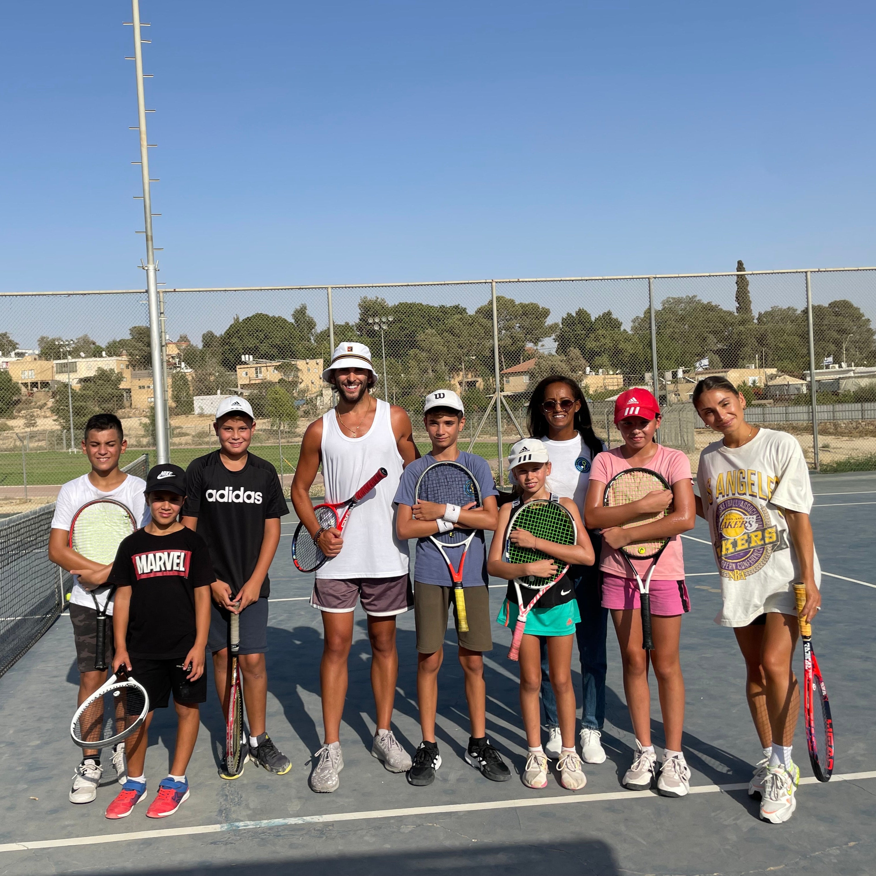 Group of people, including adults and children, standing on tennis court, holding tennis racquets